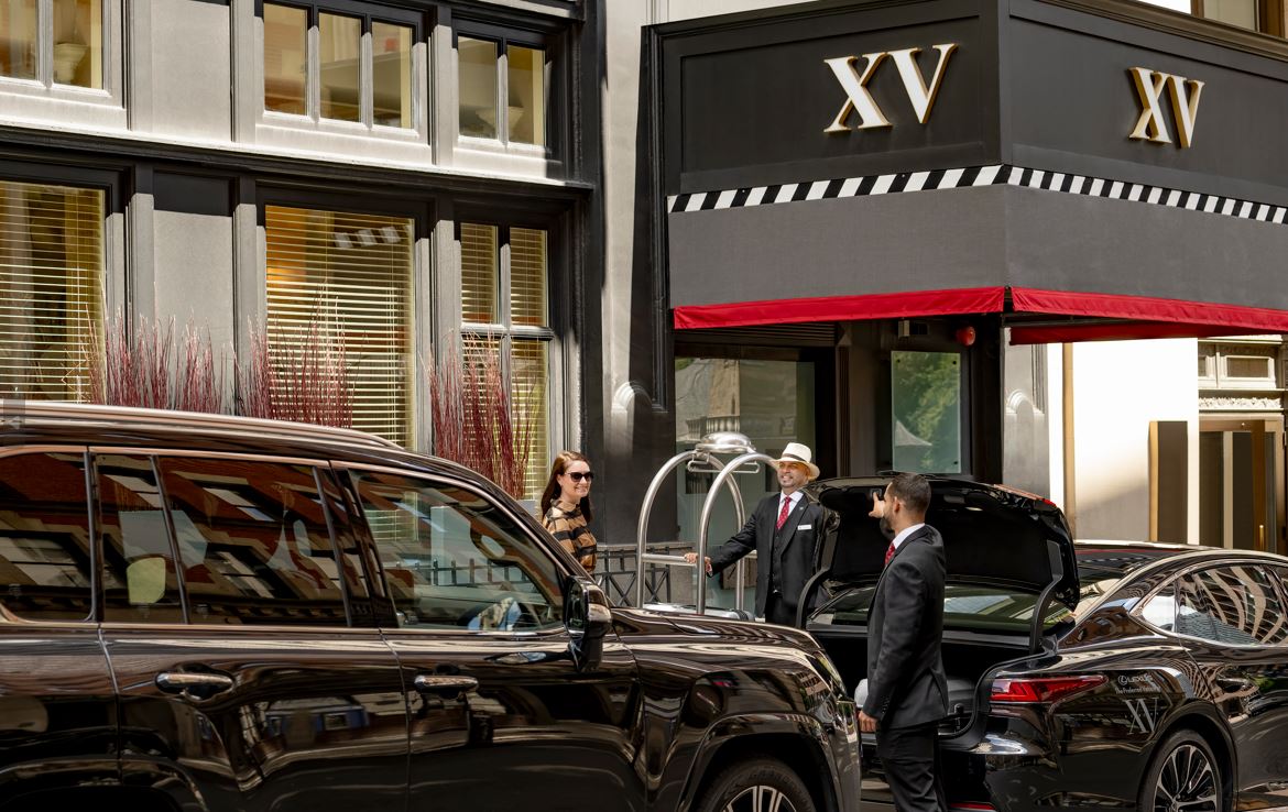 Exterior facade of the hotel. Guests are arriving in luxury vehicles. A bellman stands smiling nearby with a luggage cart.