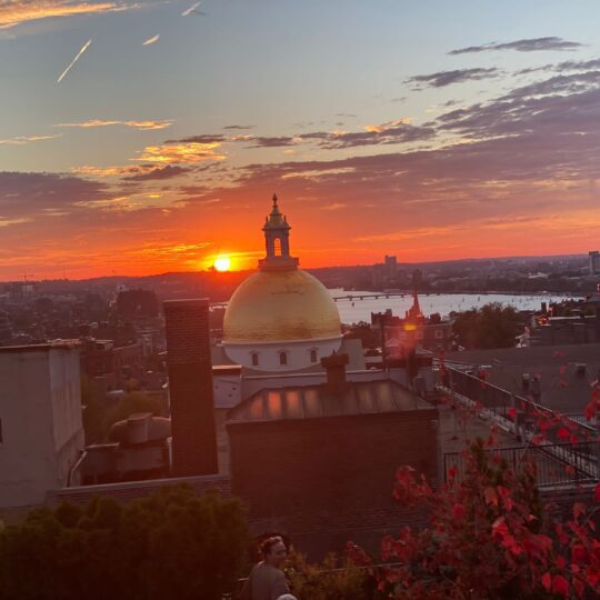 The Massachusetts State House as seen from XV Beacon hotel, with a sunset, cityscape and the Charles River in the background.